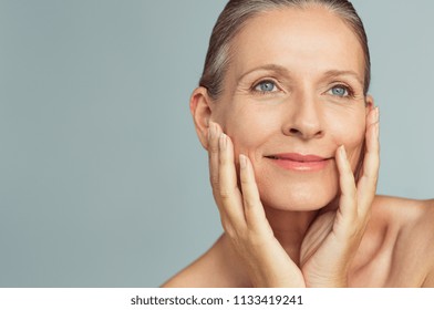 Portrait Of Mature Woman With Perfect Skin Isolated On Grey Background. Closeup Face Of Happy Senior Woman With Hands On Cheeks Looking Away. Facing Aging With A Carefree Attitude.