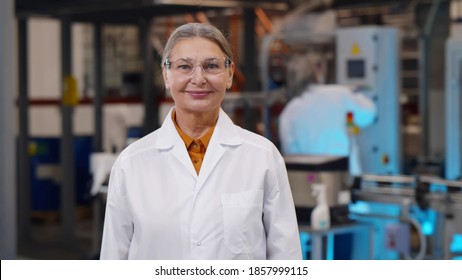 Portrait Of Mature Woman Scientist In Protective Glasses And Lab Coat Standing At Chemical Manufacturing. Aged Female Engineer Looking At Camera Over Heavy Industry Plant Background