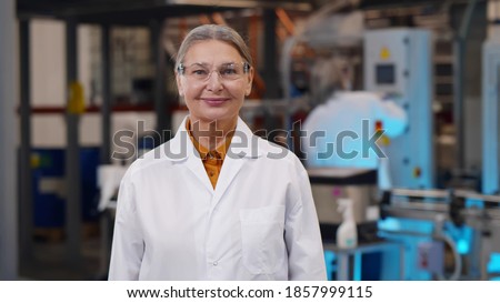Portrait of mature woman scientist in protective glasses and lab coat standing at chemical manufacturing. Aged female engineer looking at camera over heavy industry plant background