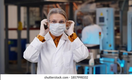 Portrait Of Mature Woman Scientist In Robe And Protective Glasses Putting On Safety Mask Standing At Plant. Chemical Manufacturing Female Engineer Wearing Safety Equipment