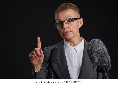 Portrait Of Mature Woman Standing At Podium And Speaking To Microphone While Giving Speech Against Black Background, Copy Space