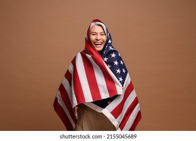 Portrait Of Mature Woman Wrapping At The USA Flag Isolated On Brown Background. Overjoyed Senior Patriotic Lady Celebrating 4th July