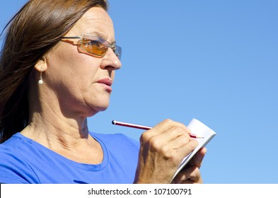 Portrait Of Mature Woman Writing Notes On Paper With Pen, Isolated With Blue Sky As Background And Copy Space.