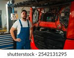 Portrait of mechanic stands confidently next to a large truck with its hood open. The mechanic wears blue overalls and a white t-shirt, with tools and equipment visible in the background.