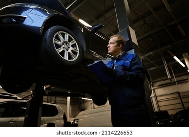 Portrait Of A Mechanic, Technician, Car Engineer In Uniform Making The Checklist On Clipboard For Repairing The Car Lifted On A Hoist In The Repair Shop During Warranty Car Maintenance. Auto Service