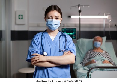 Portrait Of Medical Nurse With Chirurgical Mask In Hospital Room With Sick Unweel Senior Pantient With Trauma Looking At Camea With Arms Crossed, Wearing Scrubs.