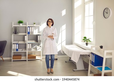 Portrait Of A Medical Worker. Friendly Woman Working As A Doctor Stands In Her Office And Looks At The Camera Smiling. Doctor In Medical Uniform, Glasses And With Tablet In Hand At His Workplace.