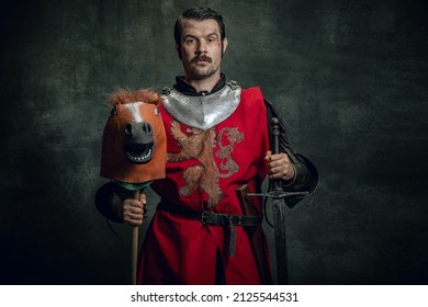 Portrait Of Medieval Warrior Or Knight Wearing Helmet And Armor Riding Toy Horse, Holding Big Sword Isolated Over Dark Background. Comparison Of Eras, History, Renaissance Style. Funny Meme