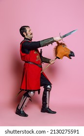 Portrait Of Medieval Warrior Or Knight Wearing Armor Clothing Riding Toy Horse, Holding Big Knife Isolated Over Pink Background. Comparison Of Eras, History, Renaissance Style. Funny Meme