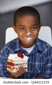 Portrait Of Messy Boy Eating Slice Of Cream Cake With Whipped Cream On Lips And Nose