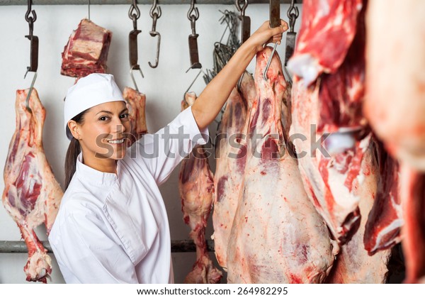 Portrait of mid adult female butcher hanging\
meat in butchery