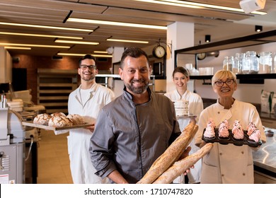 Portrait Of Mid Age Male Baker Holding Bread In His Hands At Bakery With His Multi Generation Colleagues In Backgrounds.