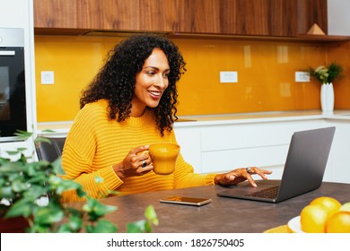 Portrait of a mid woman with black curly hair smiling while using laptop computer in kitchen  - Powered by Shutterstock