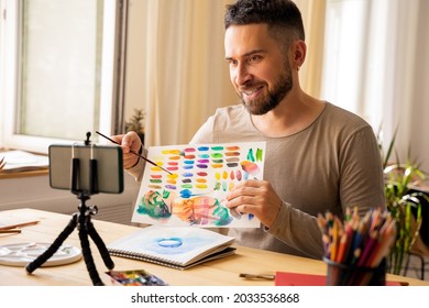 Portrait Of Mid-adult Artist Male. Dark Haired Man Smiling Happy Making Online Art Class For Students At Studio. Holding Watercolor Drawing In His Hand. Concept Zoom Remote Learning. Copy Space.