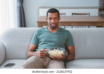 Portrait Of Middle Age African American Man Eating Popcorn While Sitting On A Couch At Home And Watching TV And Smiling At Camera. Black Man Spending Time At Home. Man Of African Ethnicity Having Fun.