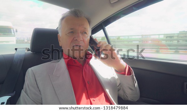 Portrait middle age businessman sitting on the\
backseat in the car. aged man sits on a seat at the back of a\
vehicle using mobile phone. Happy male in years talking on the\
smartphone drive on\
the