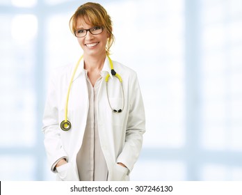 Portrait Of Middle Age Female Doctor Is Wearing A White Doctor's Coat With A Stethoscope Around Her Neck. Smiling Physician Standing At Private Clinic And Looking Away.