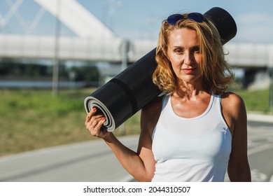 Portrait Of A Middle Age Fit Woman Holding A Rolled Yoga Mat. She Is Preparing For Hard Training On Riverside City Promenade.