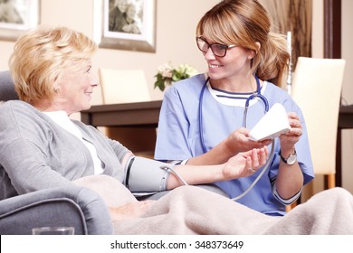 Portrait Of Middle Age Home Caregiver And Senior Woman Sitting At Nursing Home. Female Nurse Consulting With Elderly Patient While Testing Blood Pressure.