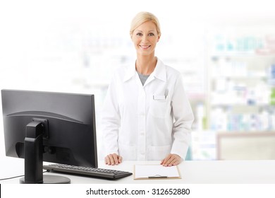 Portrait Of Middle Age Pharmacist Woman Standing At Pharmacy. Smiling Woman Wearing Lab Coat While Working On Computer And Looking At Camera. 