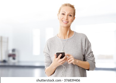 Portrait Of Middle Age Woman Using Her Mobile And Listening Music While Standing At Gym After Fitness Workout.