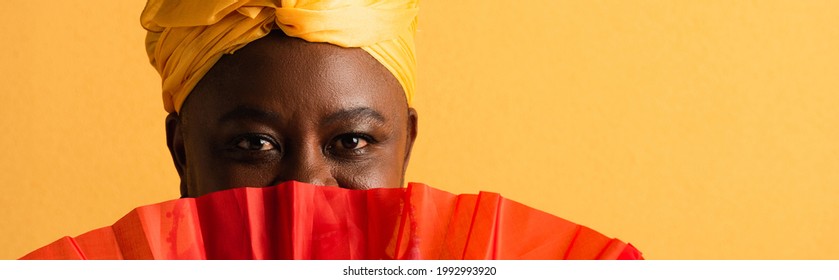 Portrait Of Middle Aged African American Woman Covering Face With Red Fan On Yellow, Banner