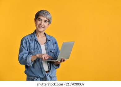 Portrait Of Middle Aged Asian Woman 50s Wearing Casual Denim Shirt White T-shirt Holding Laptop Computer And Pointing Fingers Isolated On Colour Background, Looking And Smiling At Camera