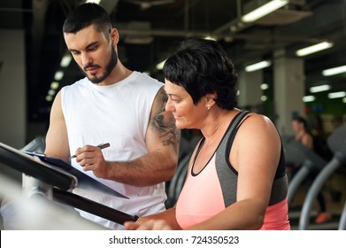 Portrait Of Middle Aged Brunette Woman Working Out With Personal Couch In Modern Fitness Center. Male Trainer And Female Client Discussing Nutrition Or Training Plan. Healthy Lifestyle Concept.