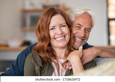 Portrait of middle aged couple hugging and looking at camera. Close up face of happy mature couple having fun at home. Senior man embracing his beautiful wife on the couch.