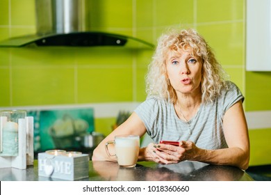 Portrait Of Middle Aged Curly Blond Caucasian Woman With Cup Of Coffee Using Online App On Her Generic Cell Phone. Serious Mature Female Speaking And Looking At Camera At Home Kitchen Interior.