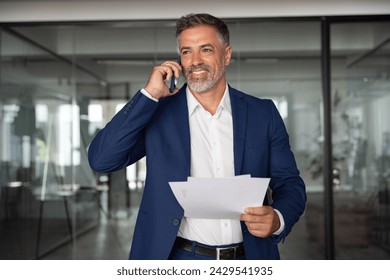 Portrait of middle aged Latin or Indian businessman walking, having call on smartphone with business partner and looking at documents. Mature Hispanic man talking by mobile cellphone at work in office - Powered by Shutterstock