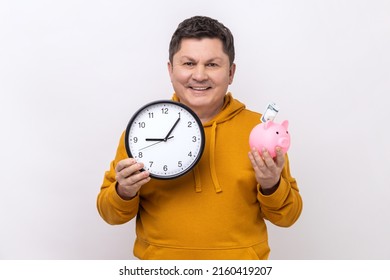 Portrait Of Middle Aged Man Holding Big Wall Clock And Piggy Bank With Banknote Inside, Looking Smiling At Camera, Wearing Urban Style Hoodie. Indoor Studio Shot Isolated On White Background.