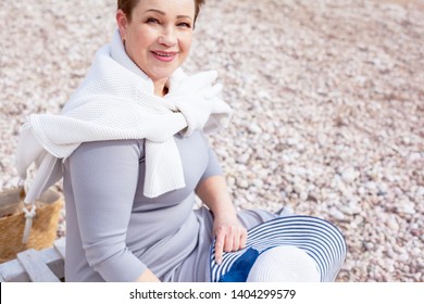 Portrait Middle Aged Mature Woman Posing On The Beach In The Sea. Wearing Grey Dress, Straw Hat And Bag