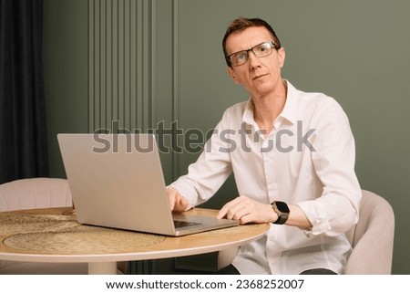 Portrait of middle aged , thin business man sitting at round table on which lying open laptop. Serious looking man in glasses and white shirt distracted from work at comfortable home.