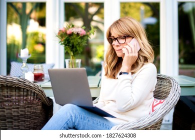 Portrait Of Middle Aged Woman Using Laptop And Talking With Somebody On Her Mobile Phone  While Working At Home In The Garden. Home Office.