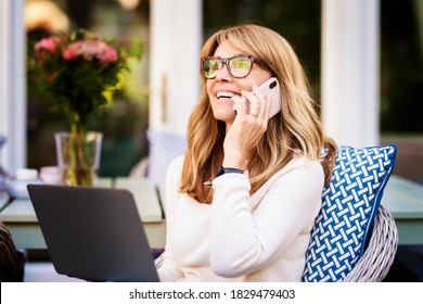 Portrait Of Middle Aged Woman Using Laptop And Talking With Somebody On Her Mobile Phone While Working At Home In The Garden. Home Office.