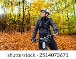 Portrait of middle aged woman wearing black sporty jacket, black cycling pants and white bike helmet standing with bicycle in forest in autumn scenery. 