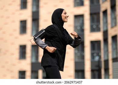 Portrait Of Middle Eastern Athletic Woman In Modest Sportswear Jogging In City, Motivated Young Muslim Lady Black In Hijab Running Near Urban Buildings, Enjoying Training Outdoors, Copy Space