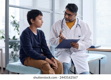 Portrait of Middle Eastern doctor talking to young boy during exam in medical clinic both sitting on cot together - Powered by Shutterstock