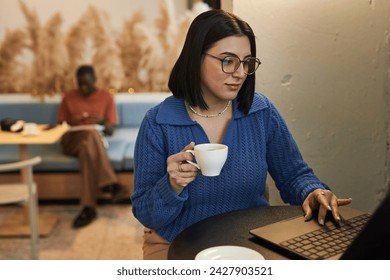 Portrait of Middle Eastern young woman wearing glasses using laptop in coffee shop and holding cup of coffee while working online copy space - Powered by Shutterstock