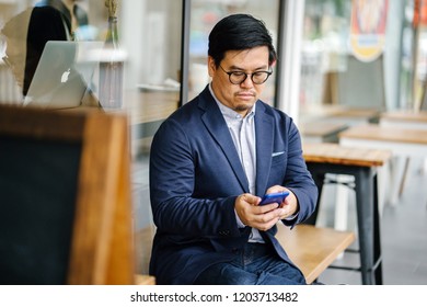 Portrait of a middle-aged Chinese Asian man in a casual suit and glasses sitting on a bench at a cafe and using his smartphone. 