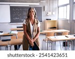 Portrait middle-aged female high school teacher looking smiling at camera with joined hands in classroom. Happy grey-haired science tutor Caucasian woman posing joyful leaning on desk in empty class 