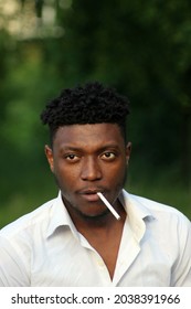 Portrait Of A Middle-aged Man With A Traditional Hairstyle. Representative Of The Race, Originally From Cameroon. African Man In A White Shirt Smokes A Cigarette.