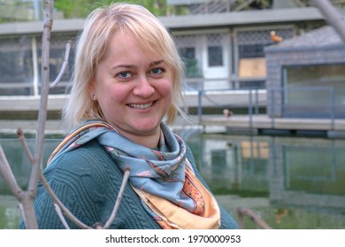 Portrait Of A Middle-aged Woman With Blond Hair Against The Backdrop Of A Pond And A Country House. Flirty Look And A Light Smile. Country House Owner, Property Manager, Casual Country Style