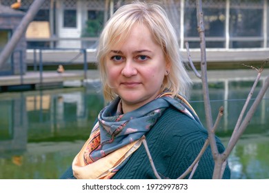 Portrait Of Middle-aged Woman With Blond Hair And Blue Eyes Against The Backdrop Of A Pond And A Country House. Woman Looking At Camera. Country House Owner, Property Manager, Casual Country Style