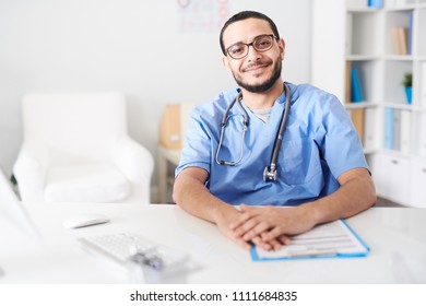 Portrait of Middle-Eastern friendly doctor wearing glasses sitting at desk in office and looking at camera smiling happily, copy space - Shutterstock ID 1111684835