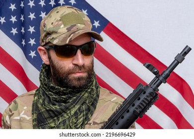 Portrait Of A Military Man 30-35 Years Old With A Rifle On His Shoulder, Wearing Dark Glasses Against The Background Of The American Flag.Concept: Military Action, Soldier Of Fortune, US Army.