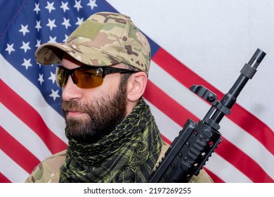 Portrait Of A Military Man 30-35 Years Old With A Rifle On His Shoulder, Wearing Dark Glasses Against The Background Of The American Flag.Concept: Military Action, Soldier Of Fortune, US Army.