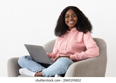 Portrait Of Millennial Black Woman Independent Contractor Sitting In Stylish Arm Chair, Using Laptop, Cheerfully Smiling At Camera, Working From Home, White Wall Background, Copy Space