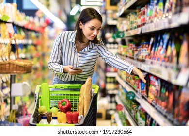 Portrait Of Millennial Lady Holding And Using Smartphone Buying Food Groceries Walking In Supermarket With Trolley Cart. Female Customer Shopping With Checklist, Taking Products From Shelf At The Shop - Shutterstock ID 1917230564
